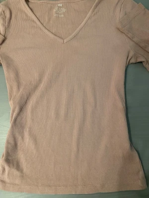 Beige ribbad topp från H&M - Säljer en beige ribbad topp från H&M med v-ringning och långa ärmar. Materialet är stretchigt och mjukt, vilket gör den superbekväm. Perfekt att styla med jeans eller kjol för en enkel och snygg look.