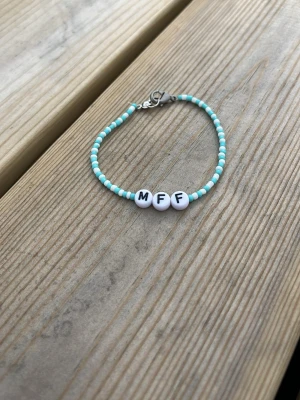 Pärlarmband med MFF-bokstäver - Snyggt armband med små turkosa och vita pärlor samt tre vita bokstavspärlor med texten 'MFF'. Armbandet har ett metallspänne och passar perfekt för dig som vill visa ditt stöd för MFF eller gillar personliga accessoarer.