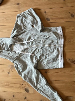 Ljusgrå hoodie från Calvin Klein - Snygg ljusgrå hoodie från Calvin Klein med dragkedja framtill och två fickor. Huva med dragsko och vit resårkant nertill med Calvin Klein-logga. Tillverkad i mjukt bomullsmaterial för en chill och bekväm stil.