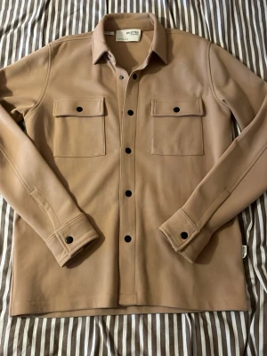 Beige overshirt från Selected Homme - Stilren beige overshirt från Selected Homme med klassisk krage, två stora bröstfickor med lock och svarta knappar framtill. Overshirten har lång ärm och är tillverkad i ett mjukt material som känns skönt mot huden. Perfekt att slänga över en t-shirt för en chill look.