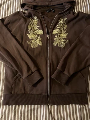 Brun hoodie med blommig brodyr - Mysig brun hoodie från ASOS DESIGN i storlek XS. Dragkedja framtill, huva och ribbade muddar. Dekorerad med snygg gul brodyr i blommönster på båda sidor av dragkedjan. Perfekt för en chill och trendig look.