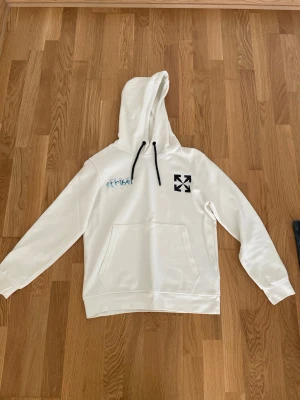 Vit hoodie från Off-White - Snygg vit hoodie från Off-White med svart logga på bröstet och coolt tryck i japansk stil på ryggen. Hoodien har svart dragsko i huvan och en stor magficka. Perfekt för dig som gillar streetwear och vill sticka ut.