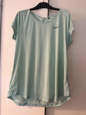 Ljusgrön sporttopp från Craft - Ljusgrön sporttopp från Craft med rund halsringning och korta ärmar. Tillverkad i ett lätt och snabbtorkande material som passar perfekt för träning. Diskret logga på bröstet och något längre baktill för extra komfort. Storlek XXL.
