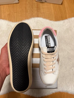 Vita GGDB sneakers med rosa detalj - Snygga vita sneakers från Golden Goose med beige stjärna på sidan och rosa detalj vid hälen. Skorna har beige snören och svart sula undertill. Klassisk låg modell i läder med GGDB-logga på plösen.