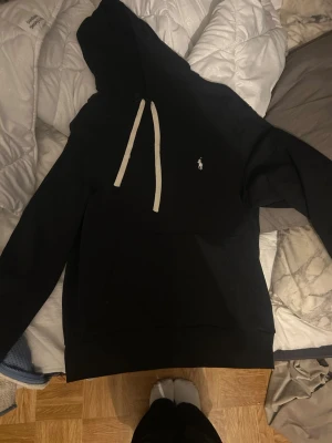 Svart hoodie från Polo Ralph Lauren - Snygg svart hoodie från Polo Ralph Lauren med vit dragsko och klassisk logga broderad på bröstet. Hoodien har en stor huva och är tillverkad i mjukt material som känns skönt mot huden. Perfekt för en avslappnad och stilren look.