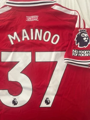 Manchester United matchtröja Mainoo - Röd Manchester United matchtröja från Adidas med Mainoo och nummer 37 på ryggen. Tröjan har vita detaljer, Premier League-märke på ärmen och ClimaCool-material som andas. Slim fit-modell med klassisk krage och klubbmärke på bröstet.