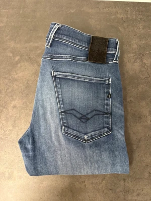 Blå slim fit jeans från Replay 33/34 - Snygga blå slim fit jeans från Replay med klassisk femficksdesign och diskret slitning. Jeansen har en ljus tvätt och detaljerad bakficka med broderad söm. Tillverkade i mjukt denimtyg för skön passform.
