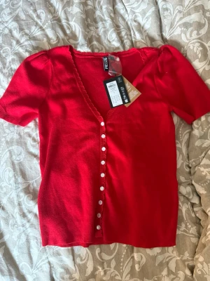 Röd v-ringad topp från Pieces - Snygg röd topp från Pieces med v-ringning och korta ärmar. Toppen har vita knappar framtill och en lätt ribbad kant längs ringningen. Perfekt för dig som vill ha en enkel men ändå trendig look.