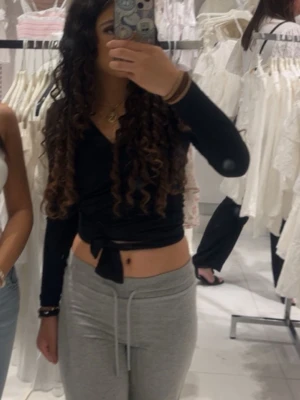 Svart omlottopp med knyt - Snygg svart omlottopp med lång ärm och knyt framtill. Toppen har en croppad passform och djup v-ringning, perfekt för att skapa en trendig look.