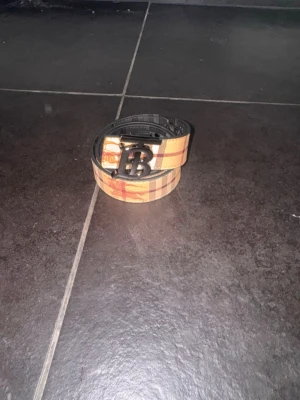 Rutig bälte med B-spänne - Snyggt bälte från med klassiskt rutigt mönster i beige, svart och rött. Bältet har ett stort svart B-spänne framtill som ger en lyxig känsla. Perfekt accessoar för att lyfta din outfit.