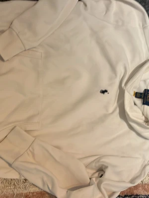 Vit hoodie från Polo Ralph Lauren - Klassisk vit hoodie från Polo Ralph Lauren med broderad marinblå logga på bröstet. Hoodien har huva, känguruficka och ribbade muddar vid ärmslut och nederkant. Tillverkad i mjukt bomullsmaterial för en skön och avslappnad look.