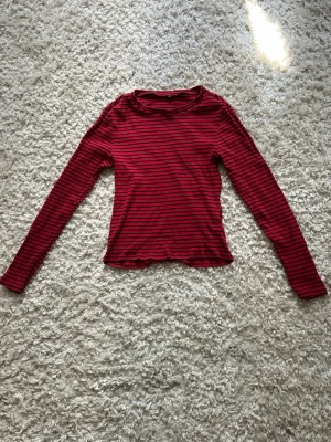 Röd och svart randig långärmad topp - En långärmad topp i rött med svarta horisontella ränder. Modellen är figurnära med rund halsringning och ribbad struktur. Perfekt att styla med jeans eller kjol för en cool vardagslook.