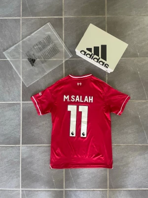 Liverpool fotbollströja Salah 11 Adidas - Röd Liverpool fotbollströja från Adidas med vita detaljer, tryck på bröstet och ärmarna samt M. Salah och nummer 11 på ryggen. Tillverkad i funktionsmaterial, perfekt för fotboll eller träning. Klassisk design med klubbmärke och sponsorlogga.