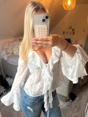 Vit volangblus från Gina Tricot - Säljer en superfin vit blus från Gina Tricot i transparent mesh med blommigt mönster. Blusen har djup urringning, volangdetaljer framtill och vid ärmslut. Perfekt för dig som gillar trendig stil.