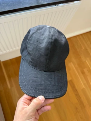 Svart keps från Fendi  - Snygg svart keps från Fendi med diskret FF-logomönster i tyget. Klassisk böjd skärm och justerbart spänne bak i läder. Perfekt accessoar för en stilren och trendig look.
