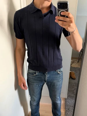 Mörkblå piké t-shirt från Dan John - Snygg mörkblå stickad t-shirt med krage och korta ärmar. Tröjan har diskreta vertikala ränder och ribbade muddar vid ärmslut och nederkant. Perfekt för en stilren och avslappnad look.