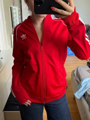 Röd Adidas ziptröja med vita ränder - Säljer en klassisk röd ziptröja från Adidas med vita ränder längs ärmarna och Adidas-logga på bröstet. Tröjan har hel dragkedja, ribbade muddar och sportig look. Perfekt för dig som gillar streetwear och vill ha en ikonisk stil❣️Tröjan är avsedd för 14-15 års i storleken, men funkar utmärkt för äldre då den är 170cm