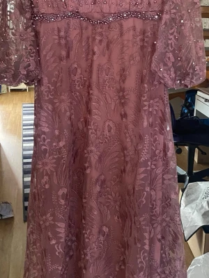 Rosa långklänning med spets och pärlor - Romantisk rosa långklänning med blommigt spetsmönster, pärldetaljer och transparenta puffärmar. Klänningen har vacker brodyr och gnistrande stenar på ärmar och överdel. Perfekt för speciella tillfällen där du vill sticka ut.passar XL , XXL