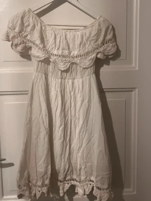 Vit offshoulder klänning med spets - Söt vit klänning med offshoulder-modell och breda spetsdetaljer längs överdelen och fållen. Klänningen har en luftig passform och är perfekt för varma dagar. Med fina virkade detaljer för en romantisk look.