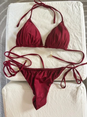 Calzedonia satin bikini - Nypris 800kr. Snygg vinröd bikini med trekantsöverdel och smala knytband både upptill och i sidorna på underdelen. Bikinin har en enkel och stilren design med små guldfärgade detaljer på snörena. 