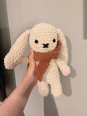 Virkad beige kanin med halsduk - Supergullig handgjord kanin i mjukt, beige garn med långa öron och svarta broderade ögon. Kaninen har en rostbrun virkad halsduk och små rosa detaljer på kinderna och tassarna. 