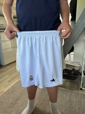 Vita Real Madrid shorts från Adidas - Säljer ett par vita Real Madrid shorts från Adidas med klubbmärket och Adidas-loggan på framsidan. Shortsen har elastisk midja och är tillverkade i ett lätt och svalt material, perfekta för fotboll eller träning.
