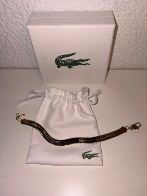 Guldigt armband från Lacoste - Snyggt guldigt armband från Lacoste med klassisk krokodil-logga ingraverad. Armbandet har en stilren och modern design med platt, ormliknande kedja och smidigt spänne. Kommer i original ask och tygpåse med Lacoste-logga. Perfekt accessoar för att lyfta din outfit.