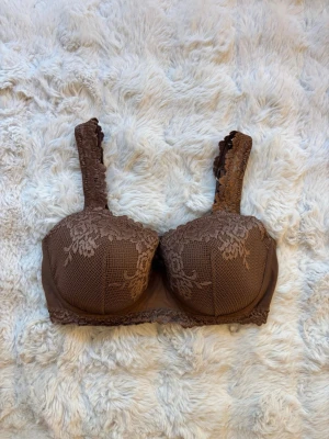 Brun Intimissimi bh med vadderade kupor - Snygg brun Intimissimi bh (Pretty flowers) med vadderade kupor och breda axelband. Dekorerad med blommig spets över hela framsidan och längs banden. Helt oanvänd. Säljer på grund av att den är lite liten.