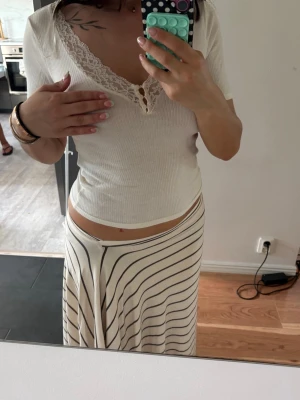 Vit ribbad topp med spetsdetalj - Säljer en söt vit ribbad topp med v-ringning och spetskant runt halsen. Toppen har korta ärmar och små knappar framtill för extra detalj. Perfekt att styla med kjol eller jeans för en fräsch look.