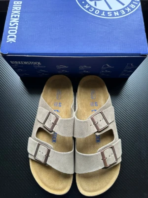 Beige Birkenstock Arizona sandaler - Säljer ett par klassiska Birkenstock Arizona sandaler i beige mocka med två breda remmar och justerbara metallspännen. Fotbädden är i kork och innersulan i mjukt material för extra komfort. Perfekta för en avslappnad och stilren look.