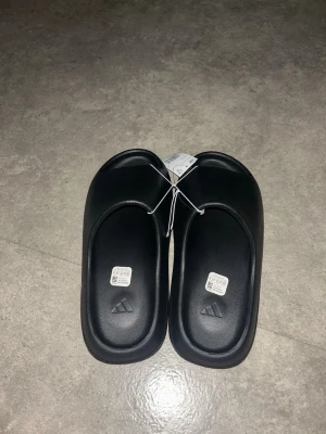 Svarta Yeezy slides från Adidas - Säljer ett par svarta tofflor från Adidas med stilren design och präglad logga på fotbädden. Tillverkade i mjukt och lätt syntetmaterial med räfflad sula för extra grepp. Perfekta för chill dagar hemma eller till och från träningen.
