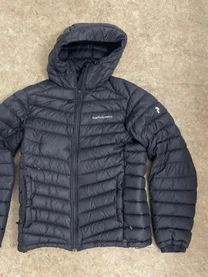 Svart dunjacka med huva från Peak Performance - Snygg svart dunjacka från Peak Performance med huva och dragkedja framtill. Jackan har två sidofickor med dragkedja och diskret logga på bröstet samt ärmen. Perfekt för kalla vinterdagar tack vare det värmande dunmaterialet.