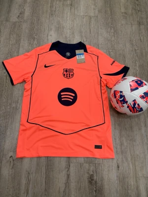 Barça Nike matchtröja neonorange - Snygg neonorange FC Barcelona matchtröja från Nike med mörkblå detaljer och loggor. Tröjan har klubbmärke, Nike-logga och Spotify som sponsor framtill samt UNHCR-logga bak. Tillverkad i lätt och ventilerande funktionsmaterial, perfekt för fotboll.