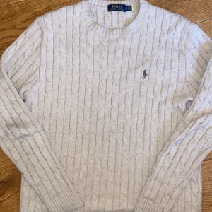 Kabelstickad ralph lauren tröja - Säljer en stilren beige stickad tröja från Ralph Lauren med kabelstickat mönster. Tröjan har långa ärmar och en rund halsringning. Jag är 185 cm lång och den sitter bra på mig. Skick 7/10 lite lös där nere men annars är den bra!