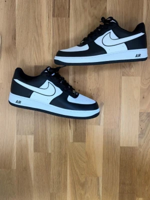Nike Air Force 1 svart/vit sneaker - Snygga Nike Air Force 1 sneakers i svart och vitt läder med klassisk vit swoosh och svart yttersula. Låga med perforerad tåbox och AIR-detalj på mellansulan. Perfekta för dig som vill ha en clean och tidlös look.