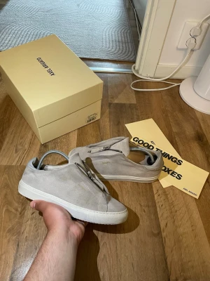 Beige sneakers från Axel Arigato - Snygga beige sneakers från Axel Arigato i mocka med dragkedja framtill istället för klassiska snören. Diskret logga på sidan och vit sula som ger en clean look. Perfekta för dig som gillar minimalistisk och modern stil. Storlek 39 passar 40