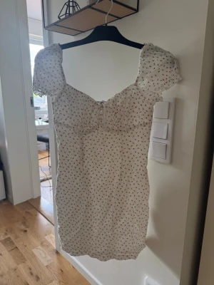 Vit blommig kortklänning Princess Polly - Supersöt vit kortklänning från Princess Polly med små röda blommor, puffärmar och söt knytning framtill. Klänningen har en feminin v-ringning och är perfekt för varma dagar. Tillverkad i mjuk bomull som känns skön mot huden. Storlek 40 men är mer en 38