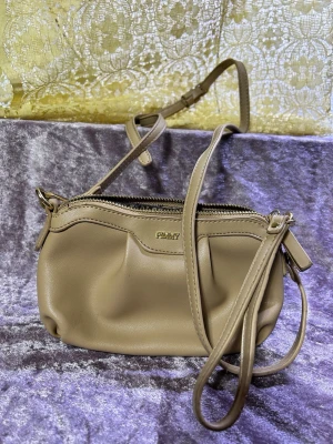 Beige axelremsväska  - Snygg beige handväska  i mjukt konstläder. Väskan har justerbar axelrem, dragkedja upptill och ett rymligt innerfack med extra ficka. Perfekt storlek för det viktigaste och stilren design med diskret logga framtill.