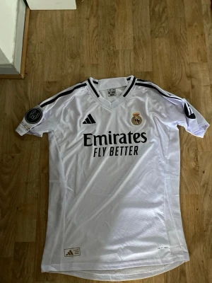 Real Madrid matchtröja Mbappé nr 9 - Säljer en vit Real Madrid matchtröja från Adidas med svarta detaljer, klubbmärke på bröstet och sponsortryck 'Emirates Fly Better'. På ryggen står det Mbappé och nummer 9. Tröjan har korta ärmar och ventilerande meshpaneler.jag Tror det är xs i storlek