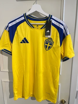 Sverige matchtröja Adidas gul/blå - Säljer en officiell Sverige matchtröja från Adidas i gult med blå detaljer och vita ränder på axlarna. Tröjan har Sveriges landslagsemblem på bröstet och Adidas-logga. Tillverkad i lätt och ventilerande polyester, perfekt för fotboll eller träning.