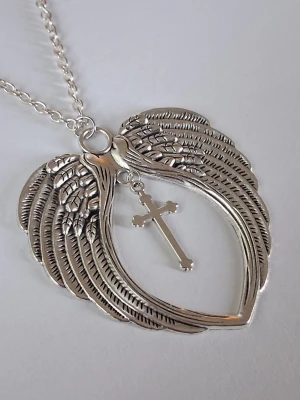 Halsband med änglavingar och kors - Snyggt halsband i silverfärgad metall med ett stort hänge format som två detaljerade änglavingar i hjärtform. I mitten hänger ett litet kors som ger en cool kontrast. unikt smycken med symbolik. Materialet rostfritt stål 
