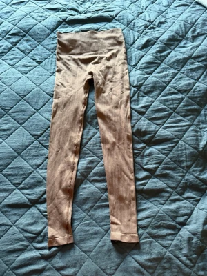 Rosa ribbade leggings - Säljer ett par beige leggings med ribbad struktur och hög midja. De är tighta och stretchiga, perfekta för en bekväm och snygg look. Materialet känns mjukt mot huden och ger bra rörelsefrihet. Inga synliga loggor eller detaljer.