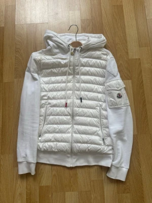 Vit jacka från Moncler - Snygg vit jacka från Moncler med quiltad dunpanel framtill och mjuka ärmar. Dragkedja hela vägen, två fickor med dragkedja och huva med dragsko. Moncler-logga på ärmen och ribbade muddar. Perfekt för dig som gillar exklusiv streetwear.