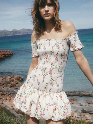  Populär Blommig offshoulder kortklänning - Söt kortklänning med offshoulder och puffig volangkjol. Klänningen är vit med blommigt mönster i rosa, gult och grönt. Smockad överdel och korta ärmar. Perfekt för varma dagar och sommarens alla tillfällen.