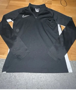 Svart långärmad träningströja Nike - Svart långärmad träningströja från Nike med vit logga på bröstet och dragkedja vid halsen. Tröjan har vita paneler längs sidorna och är tillverkad i ett mjukt, stretchigt material som passar perfekt för sport och träning. Storlek 137-147
