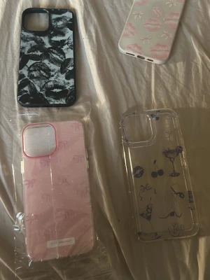 Fyrpack mobilskal i olika mönster - Säljer fyra snygga mobilskal till iPhone 13 Pro Max. Ett svartvitt med läppar, ett vitt med rosa läppar och rosetter, ett rosa med små rosetter och pärldetaljer samt ett genomskinligt med blåa drinkar och körsbär. Alla skal är i plast och har trendiga motiv. 15kr styck!!
