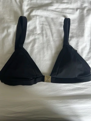 Svart bikiniöverdel med gulddetalj - Stilren svart bikiniöverdel med trekantsformade kupor och breda axelband. Snygg guldfärgad spännesdetalj framtill och justerbara band baktill för perfekt passform. Vaddering som går att ta ut. Bikinitoppen är från NA-KD och knappt använd