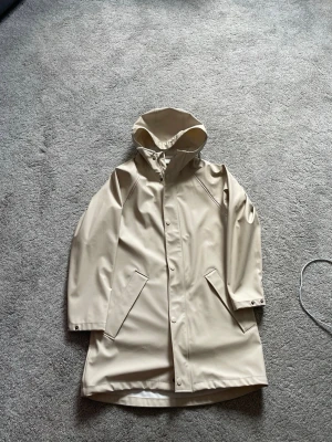 Beige regnjacka med huva - Stilren beige regnjacka med huva och tryckknappar framtill. Jackan har två snedställda fickor och är tillverkad i ett vattenavvisande material med matt finish. Perfekt för regniga dagar och enkel att matcha med olika outfits.