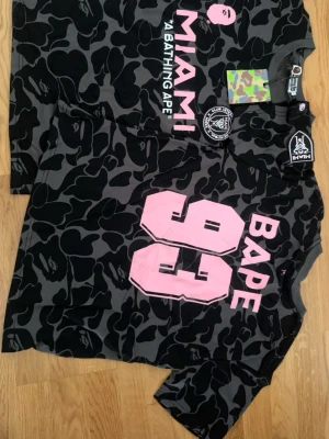 Svart camo BAPE x Miami t-shirt - Säljer en svart och grå camo t-shirt från BAPE i samarbete med Miami. T-shirten har stora rosa tryck med siffran 93 och BAPE på ryggen samt MIAMI-logga framtill. Kortärmad och tillverkad i bomull, perfekt för dig som gillar streetwear.