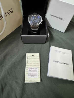 Emporio Armani silverfärgad klocka blå - Stilren klocka från Emporio Armani med silverfärgad länk i metall och mörk urtavla med flera funktioner. Klockan levereras i originalask med certifikat och manual. Perfekt accessoar för dig som vill ha en klassisk och elegant look.